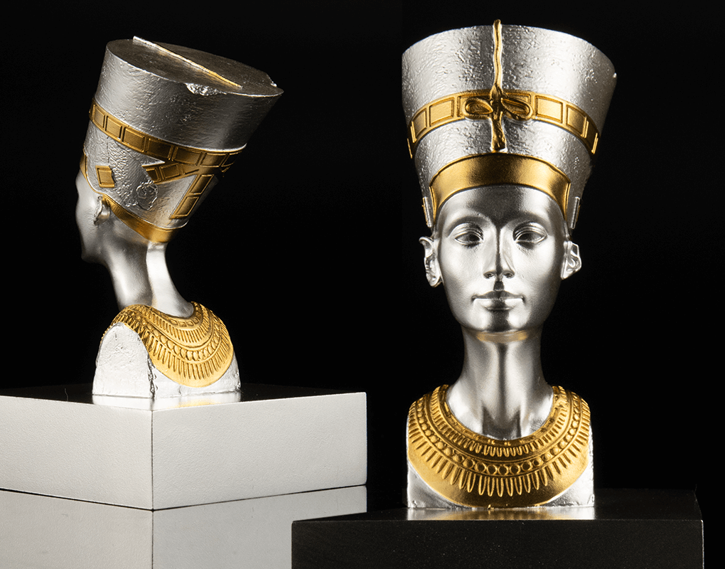 The Bust of Nefertitishowing ehind and front view of a 3D shaped silver coin with gold gilding, depicting the bust of Nefertiti. The coin is displayed on a stand against a black background.