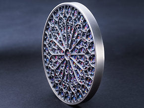 On a blue grey background the Fiji 2024 Notre Dame 5 oz Coin sits on it's rim - upright and angled A colorful mandala art design featuring intricate patterns and the Notre-Dame cathedral iconic rose window reproduction.