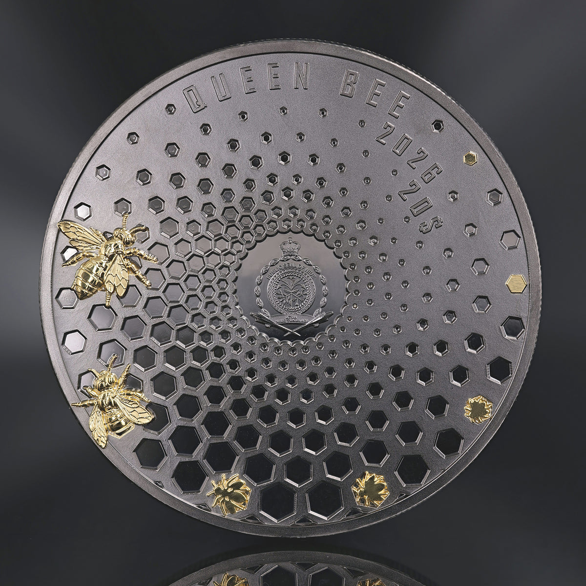 A round dark metallic coin or featuring a honeycomb pattern, embossed text reading 'QUEEN BEE' and '2026 20$', two gold-tone bees on the left, and two small gold bees birthing from the comb. Coin is photographed on a dark greyreflective background.