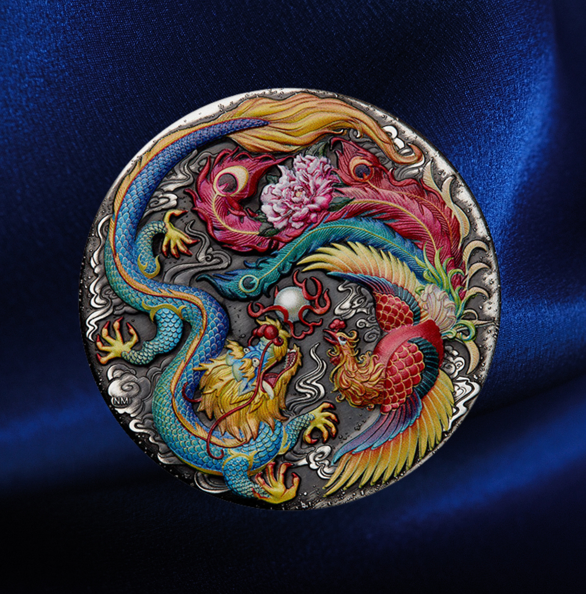 A circular silver-toned metal medallion featuring a highly detailed, colorful Chinese dragon and phoenix design, displayed on a draped dark blue satin fabric background. The coin is a round silver-toned metal medallion featuring a detailed relief design of a blue dragon and a red phoenix surrounding a pink peony, set against a dark blue satin background.