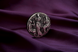 A silver coin with a detailed relief design featuring three ancient goddesses, Clotho, Lachesis, and Atropos, from Greek mythology, within a grove setting. The Coin is in a setting of luxurious purle velvet.