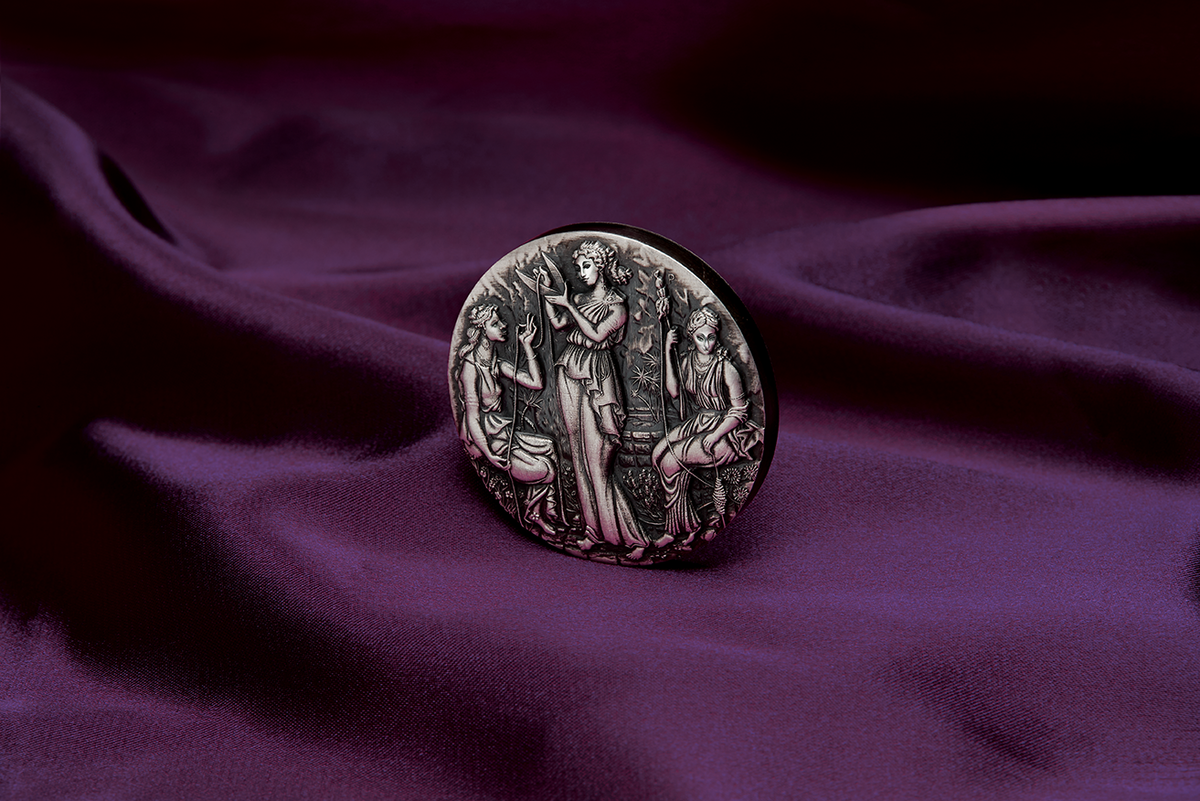 A silver coin with a detailed relief design featuring three ancient goddesses, Clotho, Lachesis, and Atropos, from Greek mythology, within a grove setting. The Coin is in a setting of luxurious purle velvet and tilted t face the left.
