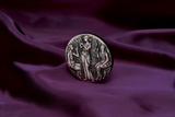 A silver coin with a detailed relief design featuring three ancient goddesses, Clotho, Lachesis, and Atropos, from Greek mythology, within a grove setting. The Coin is in a setting of luxurious purle velvet and tilted t face the left.
