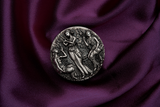 A silver coin with a detailed relief design featuring three ancient goddesses, Clotho, Lachesis, and Atropos, from Greek mythology, within a grove setting. The Coin is in a setting of luxurious purle velvet lying flay giving a birdseye view. The coin is rotated to 11 o'clock.
