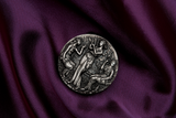 A birdseye view of a silver coin with a detailed relief design featuring three ancient goddesses, Clotho, Lachesis, and Atropos, from Greek mythology, within a grove setting. The Coin is in a setting of luxurious purle velvet. The coin is rotated 1 hou to 1 o'clock.