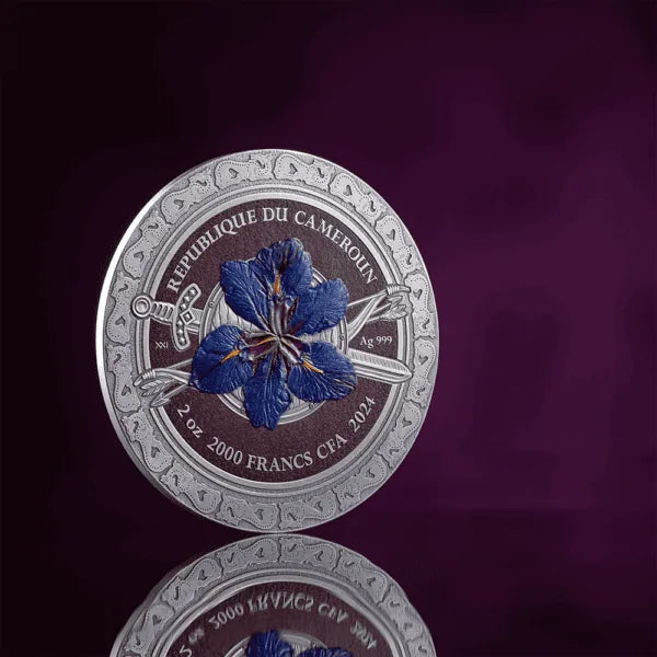 A circular silver-toned commemorative coin from the Republic of the Cameroan central bank, featuring a blue iris flower in the center and French text around the edge, shown on a dark purple reflective background.
