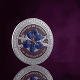 A circular silver-toned commemorative coin from the Republic of the Cameroan central bank, featuring a blue iris flower in the center and French text around the edge, shown on a dark purple reflective background.