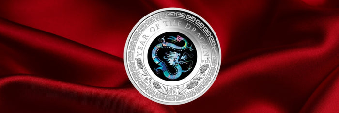 A round silver-colored collectible coin featuring a detailed blue and black dragon design in the center, surrounded by the text 'YEAR OF THE DRAGON'. The coin is displayed on a draped, shiny red satin satin fabric background.