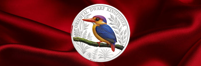 A circular silver coin featuring a colorful bird perched on a branch, set against a draped red satin background. The coin is inscribed with 'ORIENTAL DWARF KINGFISH' and shows a blue, purple, orange, and red bird.