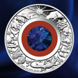 A round silver coin featuring detailed animal and celestial motifs around the border, with a central blue faceted gemstone and concentric red, white, and silver rings. The coin is photographed against a dark blue textured background.