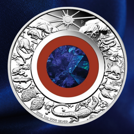 A round silver coin featuring detailed animal and celestial motifs around the border, with a central blue faceted gemstone and concentric red, white, and silver rings. The coin is photographed against a dark blue textured background.