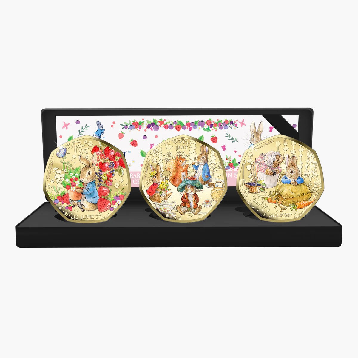 A set of three round gold-toned medallion coins featuring illustrated rabbits from Peter Rabbit, displayed on a black stand with a floral insert card behind them.