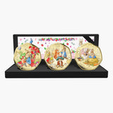 A set of three round gold-toned medallion coins featuring illustrated rabbits from Peter Rabbit, displayed on a black stand with a floral insert card behind them.