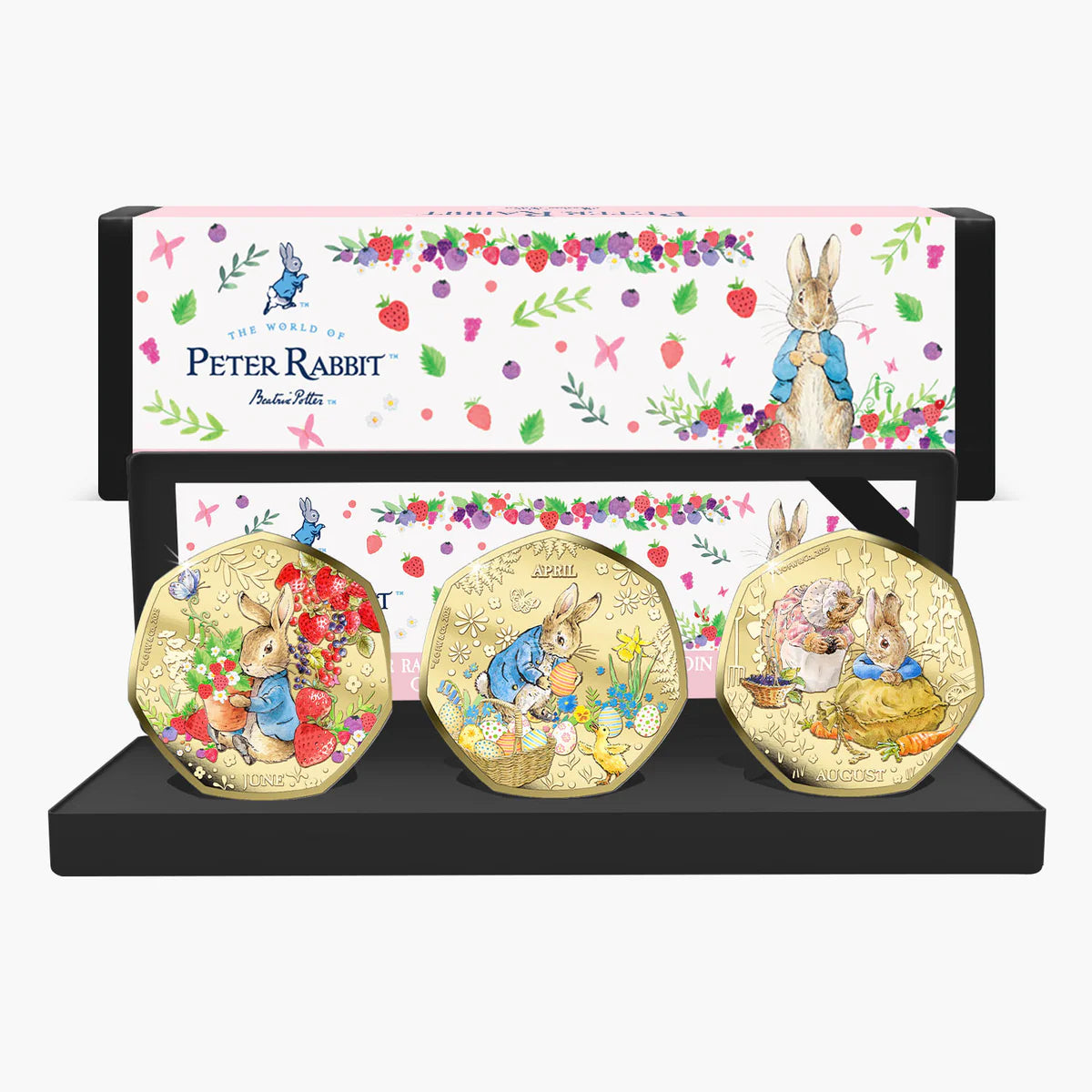 Peter Rabbit 3-piece gold coin set displayed on a black stand in front of its illustrated gift box. The coins feature Beatrix Potter characters in colorful spring scenes, and the box is decorated with Peter Rabbit illustrations and floral patterns.