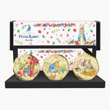 Peter Rabbit 3-piece gold coin set displayed on a black stand in front of its illustrated gift box. The coins feature Beatrix Potter characters in colorful spring scenes, and the box is decorated with Peter Rabbit illustrations and floral patterns.