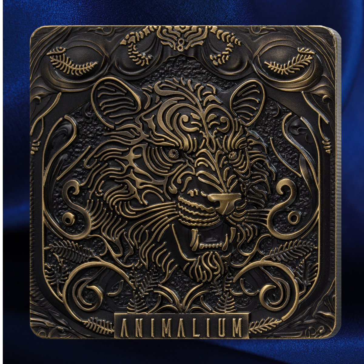 A square decorative plaque or box cover featuring an ornate embossed lion head in metallic gold against a dark bronze background, with the word 'ANIMALIUM' at the bottom, set against a blue fabric backdrop.