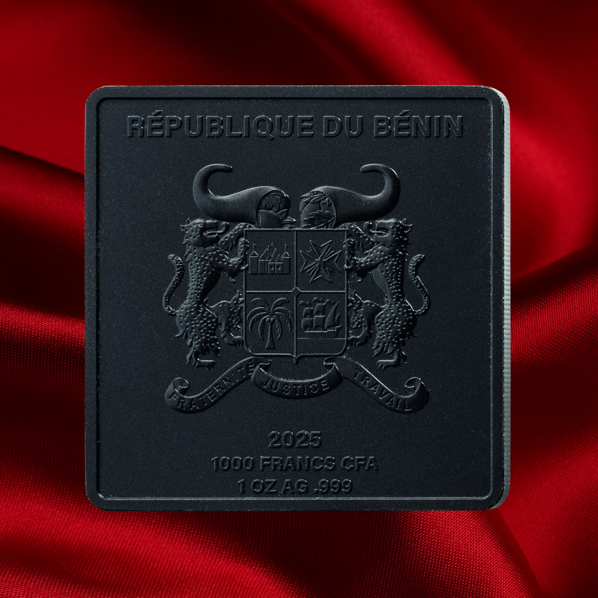 A square black commemorative coin or medallion embossed with the Republic of Benin coat of arms and text reading '2025 1000 FRANCS CFA 1 OZ AG .999', photographed against draped red satin fabric.