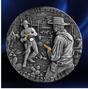 A round silver-toned collectible coin or medallion featuring a detailed relief design of a Wild West cowboy duel scene, with two cowboys facing each other holding pistols and the words 'WILD WEST' at the top and 'COWBOY DUEL' at the bottom. The coin is photographed against a dark blue satin-like background.