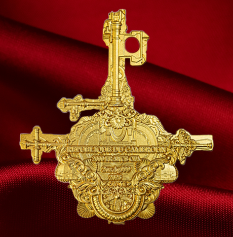 A gold-colored, highly ornate collectible pin or medallion featuring crossed keys and decorative scrollwork, displayed against a draped red satin background.