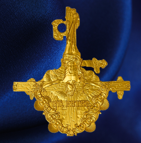 A gold-toned ornate religious pendant or badge featuring a raised figure and decorative scrollwork, shown against a dark blue satin-like background.