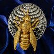 A gold bee figurine positioned in front of a silver beehive-shaped decorative object, set against a dark blue velvet fabric background.