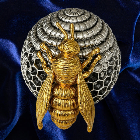 A gold bee figurine positioned in front of a silver beehive-shaped decorative object, set against a dark blue velvet fabric background.
