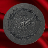 A round dark metallic coin or medallion with embossed text reading 'REPUBLIQUE DU CAMEROUN', '2025', and '3000 FRANCS CFA AG 999 3 OZ', centered on a rich red satin fabric background.