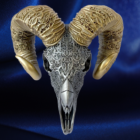 An ornate ram skull wall hanging or sculpture with large curved horns, featuring intricate filigree and engraved detailing in silver and gold tones against a dark blue satin background.