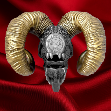 An ornate ram skull bust with large curved golden horns and a silver-toned skull center featuring a circular emblem, displayed against a rich red satin background.