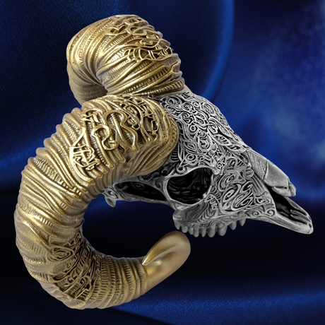 A gothic-style ring shaped like a ram skull with one horn made of textured gold-toned metal and the other half made of intricate silver-toned filigree, set against a dark blue satin background.