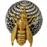 A gold bee figurine positioned in front of a silver beehive-shaped decorative object, set against a dark blue velvet fabric background.