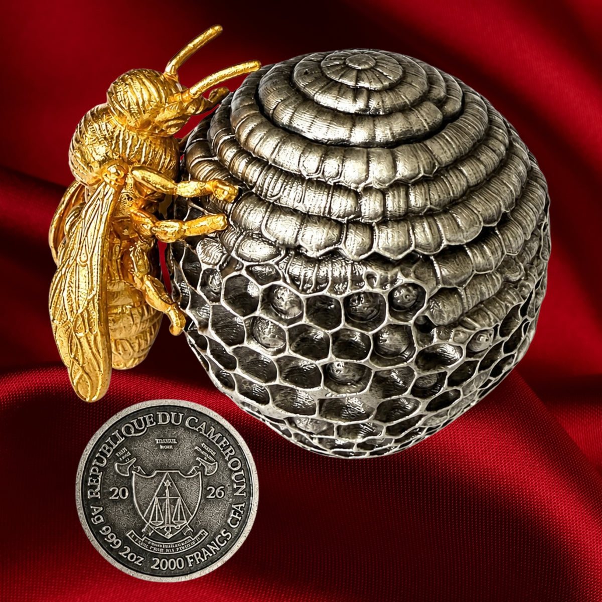 A detailed silver beehive- with a gold bee perched on the left side, displayed on a draped red fabric background the obverse of the coin which is the base of the Beehive is shown below the bee.