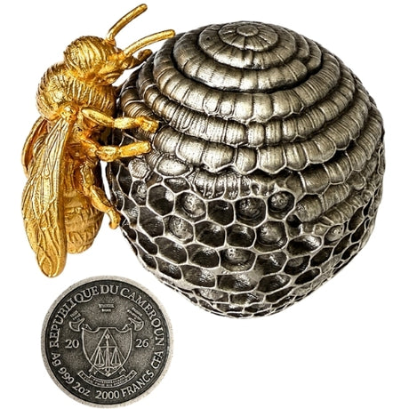 A detailed silver beehive- with a gold bee perched on the left side, displayed on a draped red fabric background the obverse of the coin which is the base of the Beehive is shown below the bee.