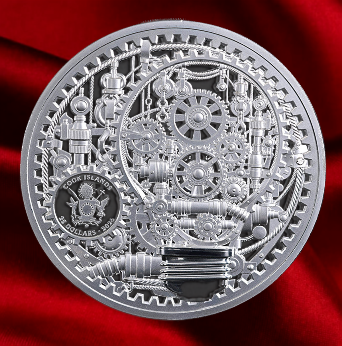 A circular silver collectible coin featuring intricate steampunk gear and cogs, with a black inset showing the Cook Islands emblem and text. The coin is photographed on a rich red satin fabric background.