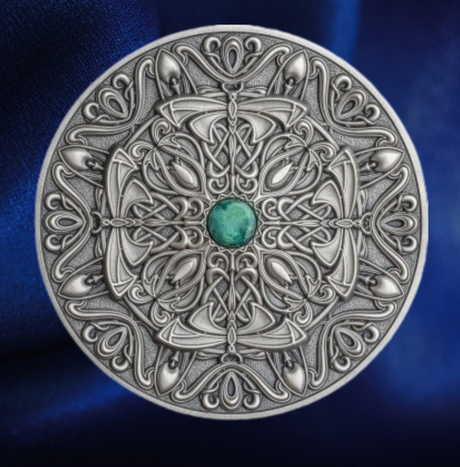 An ornate round silver-toned coin  featuring intricate Celtic knotwork and scrollwork, with a small green gemstone at the center, set against a dark blue satin background.