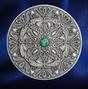 An ornate round silver-toned coin  featuring intricate Celtic knotwork and scrollwork, with a small green gemstone at the center, set against a dark blue satin background.