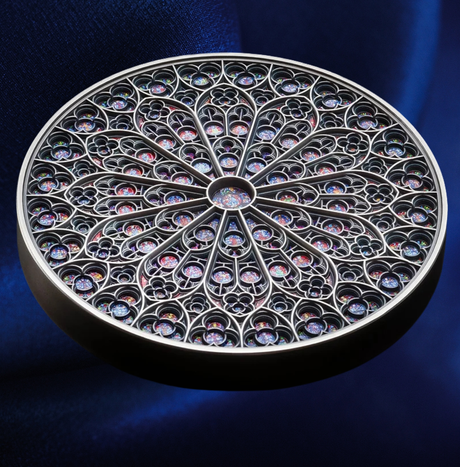 A circular silver-toned filigree jewelry box or compact with an intricate rose-meadow pattern of small iridescent gemstone compartments, set against a dark blue velvet background.