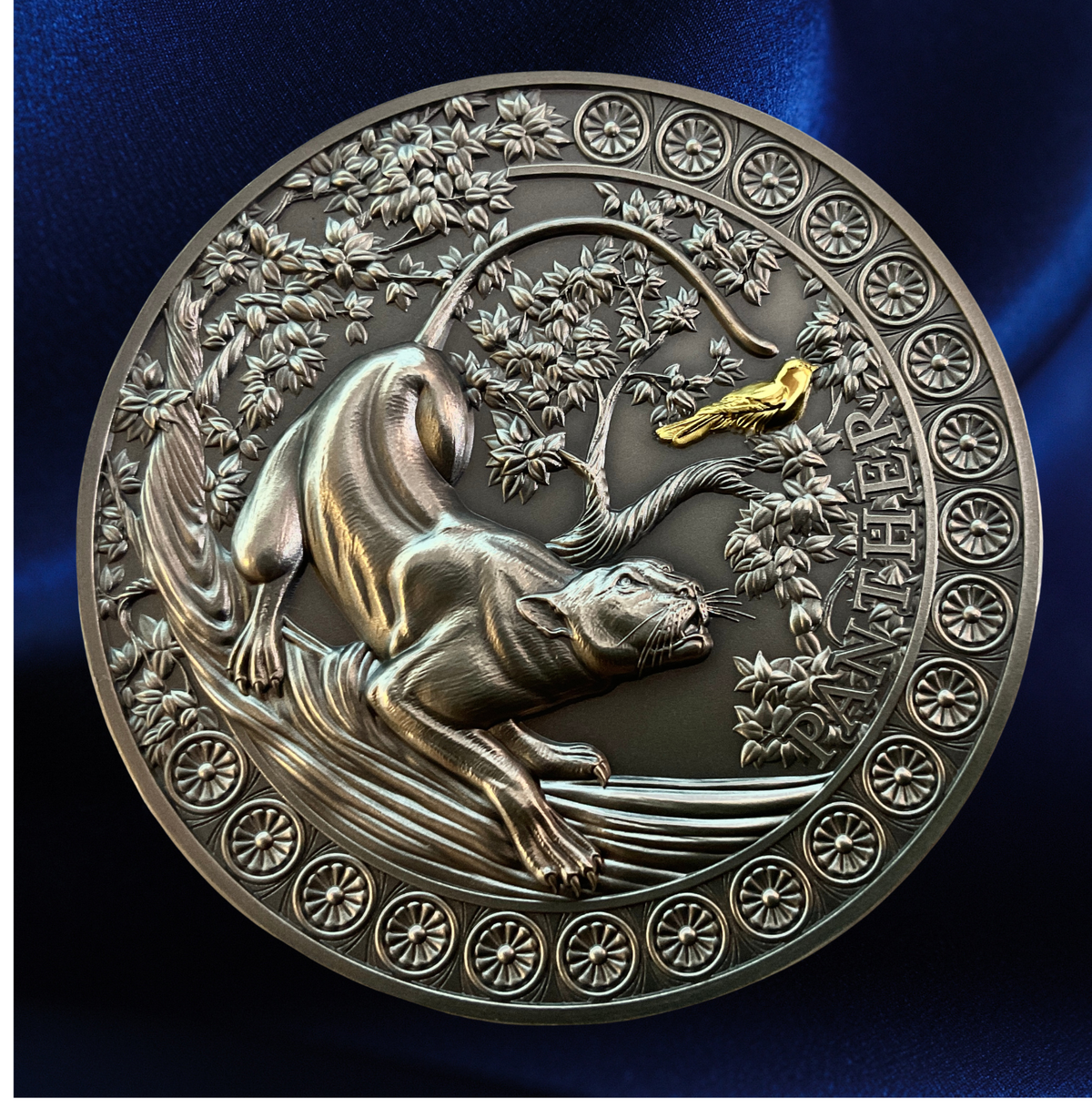 A round metallic medallion or coin featuring an embossed animal and floral design, with a small gold accent near the top right, shown against a dark blue satin-like background.
