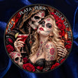 A circular collectible coin or medallion featuring a Day of the Dead theme with a hooded skeleton figure and a tattooed woman surrounded by red roses and a decorated skull, set against a dark blue satin fabric background.