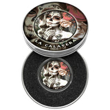 A round silver-toned metal tin shown open, displaying a Day of the Dead La Calavera coin. The same coin artwork is on the lid and inside a black foam insert. The artwork features a sugar-skull woman with a rose and decorative text.
