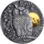 A high relief design of a long-eared owl perched above a gilded moon on a silver coin, with leaves and branches in the background.