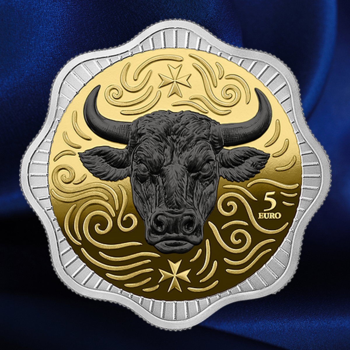 A 5 Euro coin featuring a black bull head on a gold background with decorative swirls and a silver-toned outer border, shown against a dark blue satin-like backdrop.