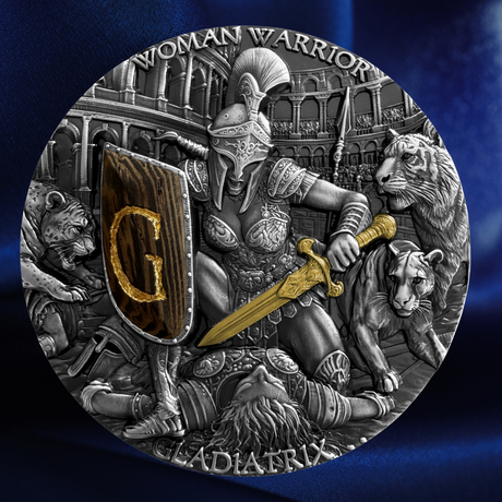 A circular silver-toned coin or medallion featuring a detailed gladiator scene with a warrior holding a sword and shield marked with a gold letter G, surrounded by animals and an arena background. The text 'WOMAN WARRIOR' and 'GLADIATRIX' is visible around the border.
