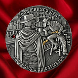 A circular silver-toned commemorative coin or medallion featuring two illustrated bar patrons with gilded guns aimed at each other, with raised text reading '2000 FRANCS - CFA and REPUBLIQUE DU CAMEROUN