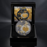 A round collectible coin featuring gold and silver bee and honeycomb designs, displayed inside an open black presentation case with a printed certificate of Authenticity.