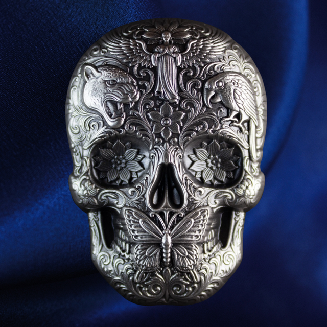 An ornate silver-toned skull pendant or decorative object with intricate floral, bird, and butterfly engravings, shown against a deep blue fabric background.