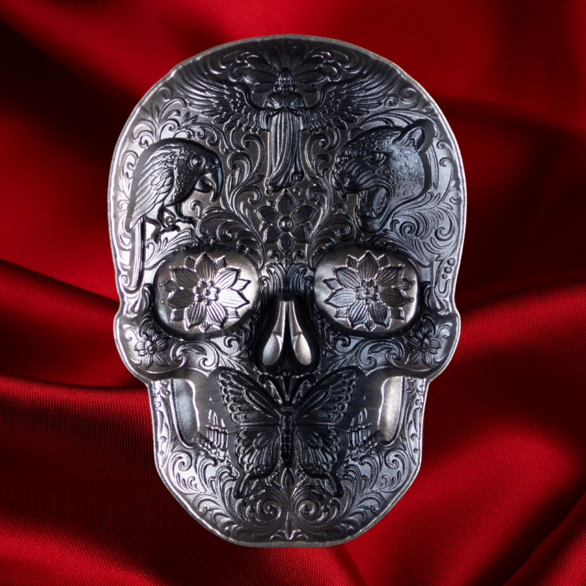 An ornate metallic skull-shaped object with intricate embossed floral and animal motifs, photographed against draped red fabric.
