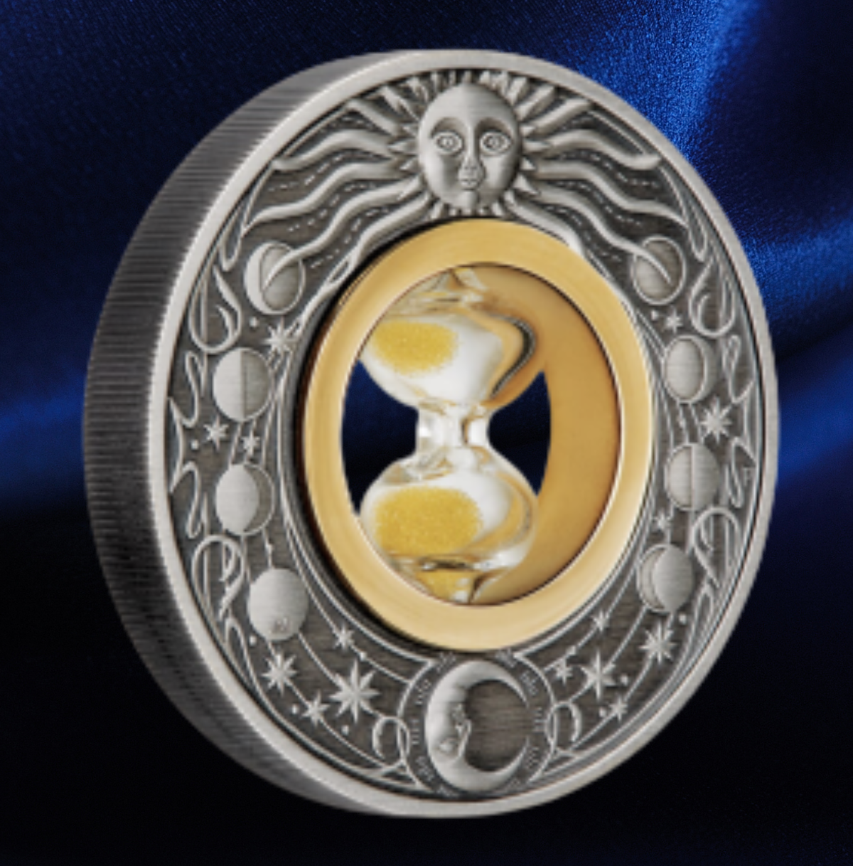 A round silver-toned medallion or coin featuring a sun face at the top, moon phases around the border, and a central gold-toned oval with an hourglass. The piece is photographed on a dark blue satin-like background.