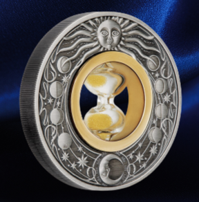 A round silver-toned medallion or coin featuring a sun face at the top, moon phases around the border, and a central gold-toned oval with an hourglass. The piece is photographed on a dark blue satin-like background.