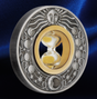 A round silver-toned medallion or coin featuring a sun face at the top, moon phases around the border, and a central gold-toned oval with an hourglass. The piece is photographed on a dark blue satin-like background.